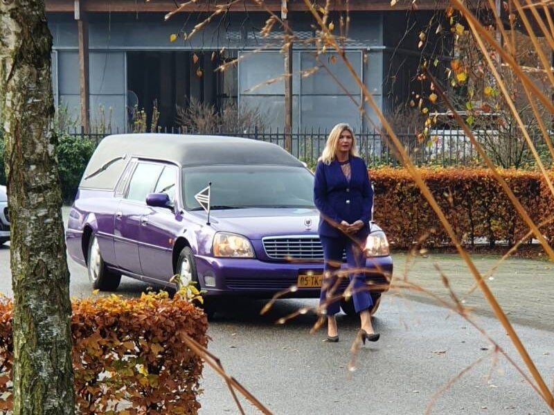 Begrafenisondernemr in Wageningen loopt voor een paarse rouwauto in paarse kleding in een herfstachtige omgeving