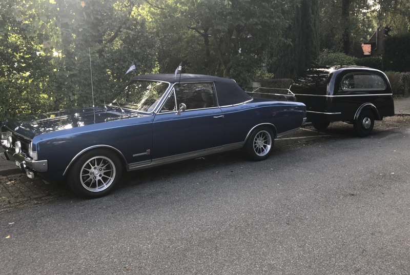 Blauwe klassieke cabriolet met een zwarte uitvaartaanhanger, geparkeerd in een groene en bosrijke omgeving.