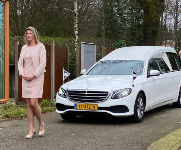 Uitvaartverzorger in roze kleding loopt voor een witte rouwwagen