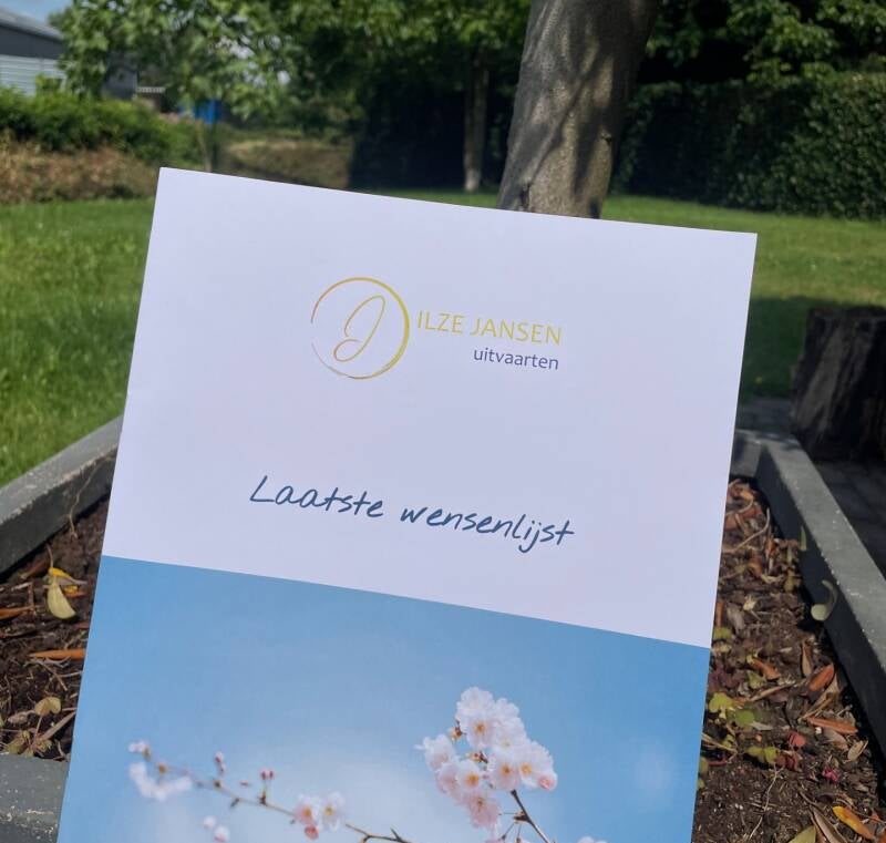 Een foto van een 'Laatste wensenlijst' brochure van Ilze Jansen Uitvaarten, met een bloemmotief en een groene tuin op de achtergrond