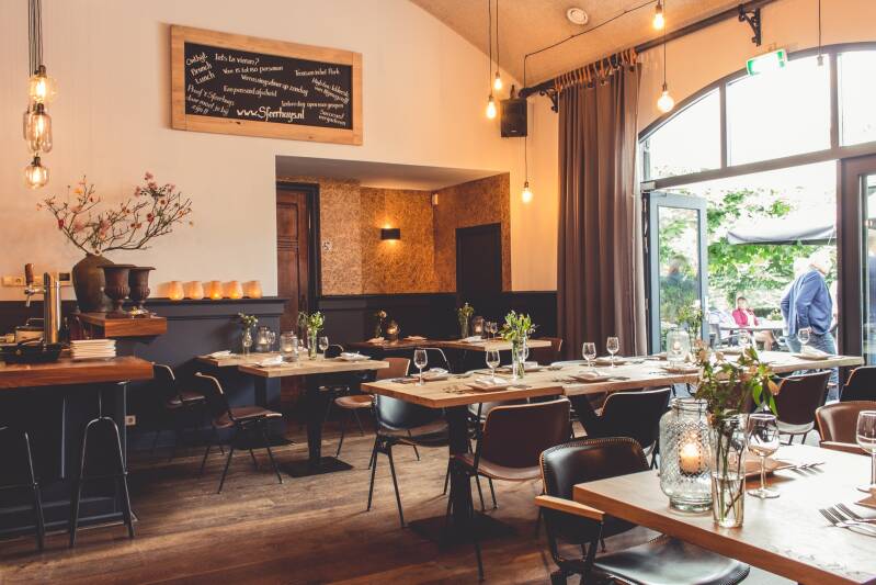 Een sfeervolle uitvaartlocatie of evenementenruimte met gedekte tafels, kaarsen en een chique, rustige inrichting. De ruimte heeft een open deur naar een buitenruimte met gasten.