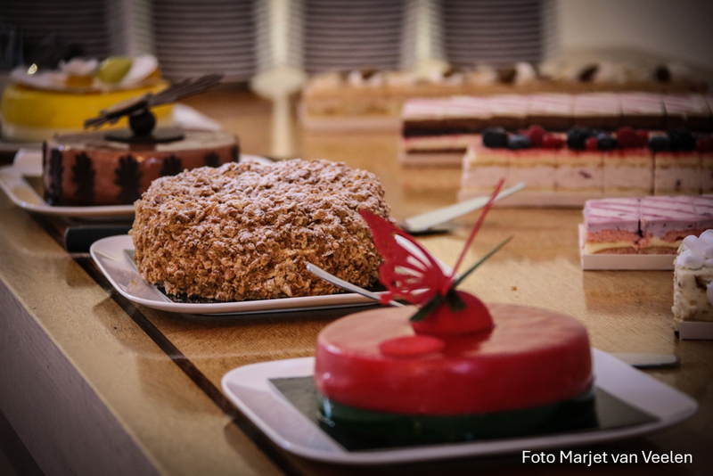 Assortiment van diverse taarten en gebakjes op een houten tafel, waaronder een rode taart met decoratieve vlinder, een kruimeltaart en verschillende lagengebak met fruit.