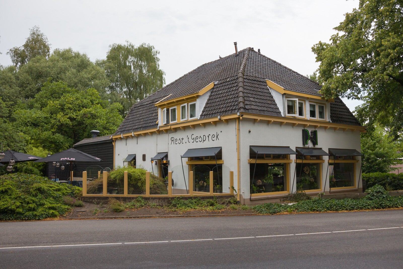 Het pand van restaurant 't Gesprek met een karakteristieke gevel, terrasstoelen en bomen rondom, gelegen aan de straat