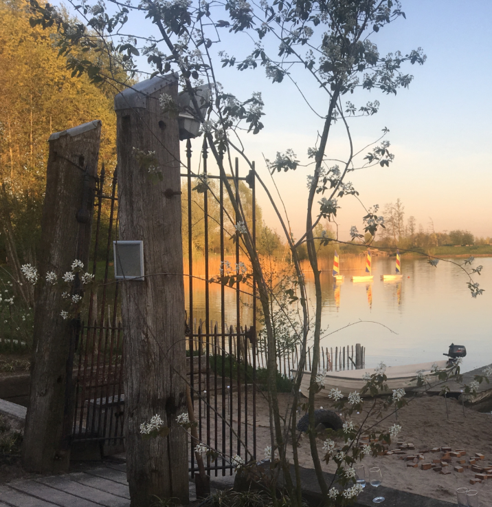 Rustieke houten poort aan het water bij zonsondergang, met doorkijk naar een rustig meer en kleurrijke zeilbootjes – ideale natuurrijke locatie voor een intiem afscheid of ceremonie in de buitenlucht.