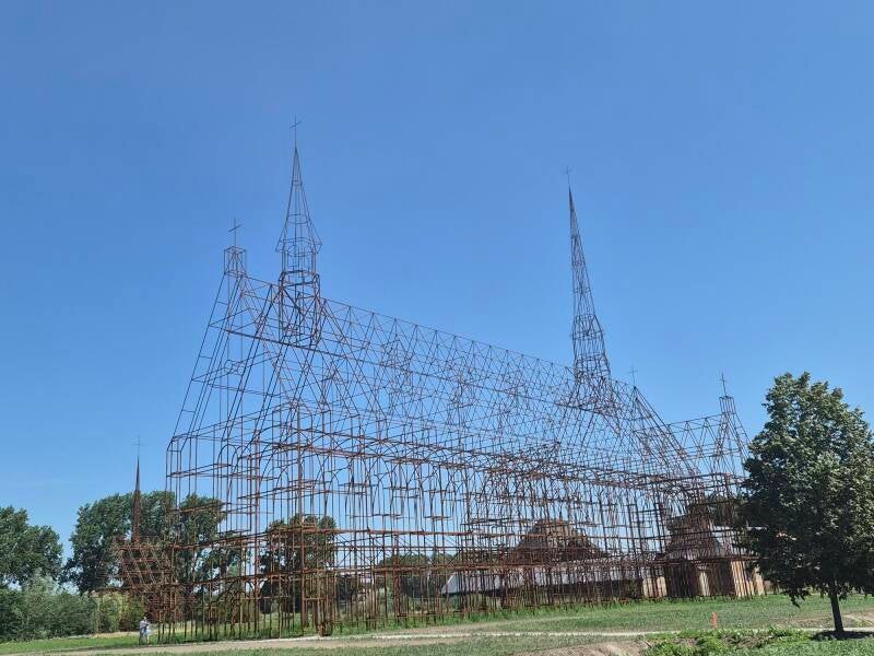 Een reusachtige, opengewerkte metaalconstructie van cortenstaal in de vorm van een historische kerk met twee spitse torens. De constructie staat in een open, grasrijk landschap onder een strakblauwe lucht