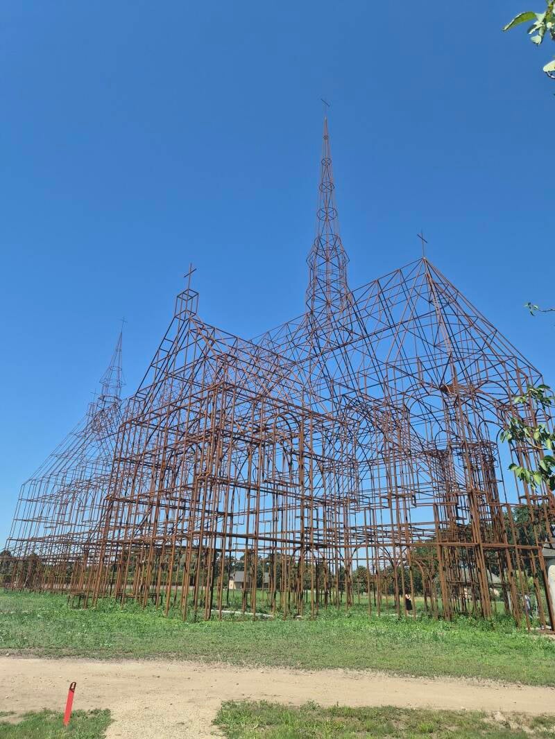Een reusachtige, opengewerkte metaalconstructie van cortenstaal in de vorm van een historische kerk met twee spitse torens. De constructie staat in een open, grasrijk landschap onder een strakblauwe lucht