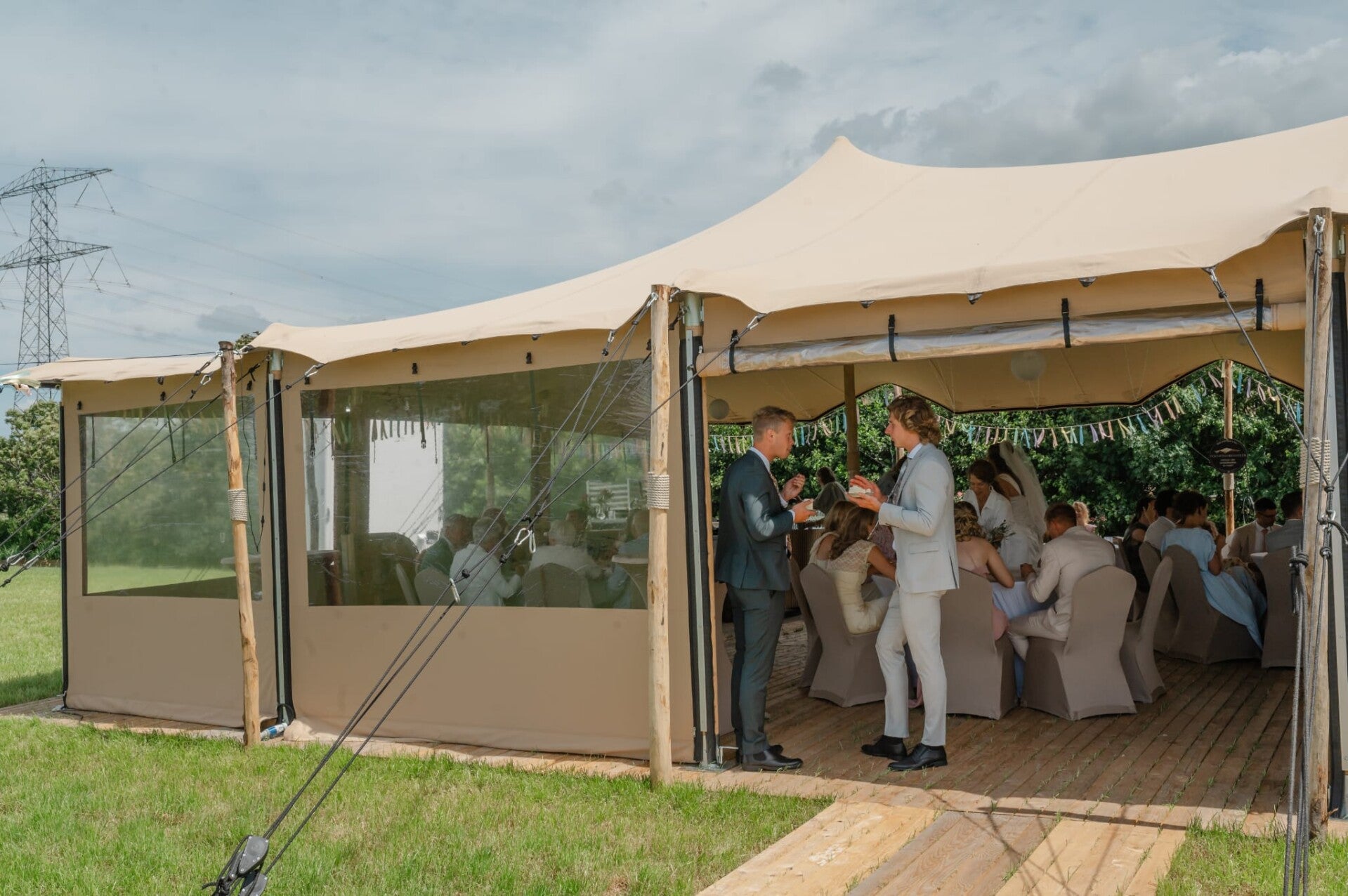 Feestgasten onder een zandkleurige stretchtent met transparante zijwanden en een houten vlondervloer