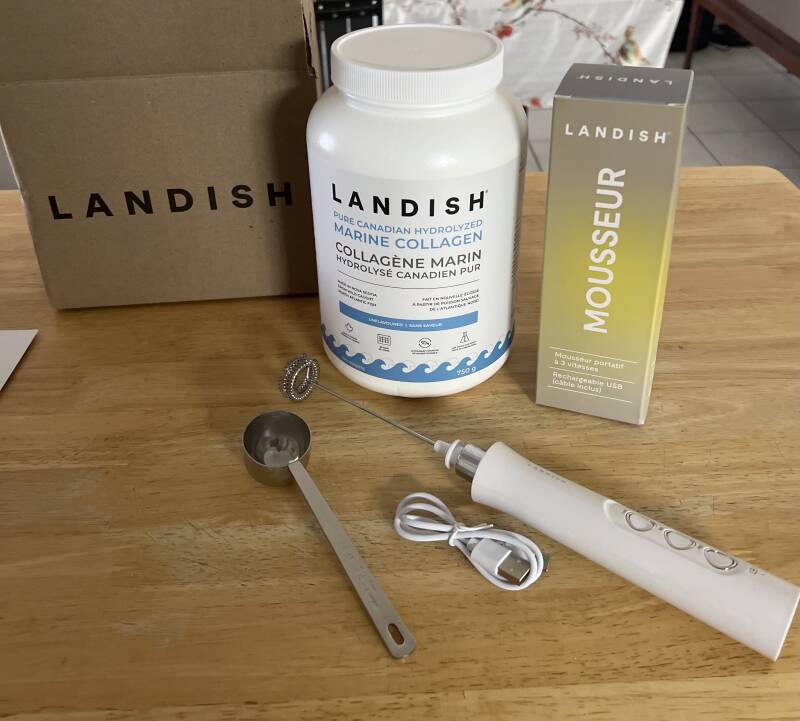 Un pot de collagène marin hydrolysé canadien pur Landish posé sur une table en bois à côté de sa boîte d'expédition, d'un mousseur à lait portatif blanc avec son câble de recharge USB, d'une boîte d'emballage marquée Mousseur et d'une cuillère à mesurer..