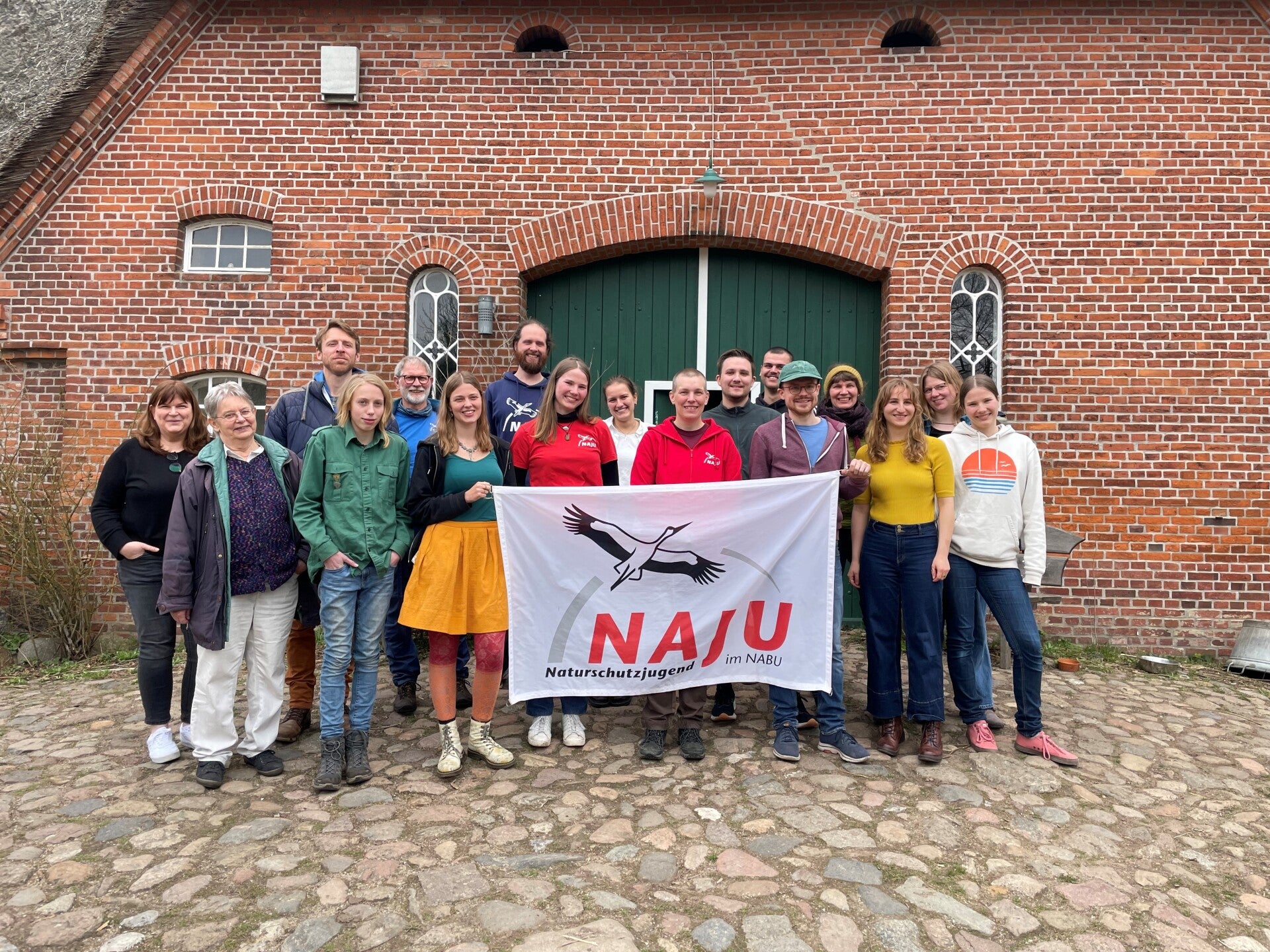 Gruppe von Menschen steht vor einem historischen Backsteingebäude und hält ein NAJU-Banner für ein gemeinsames Gruppenfoto.