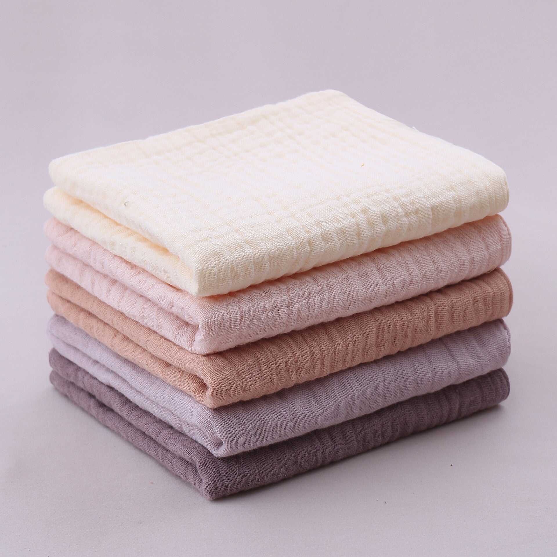 A neat stack of five folded organic cotton muslin squares from The Little Soft Cloth Lavender Fields Collection, showcasing a soft color gradient from top to bottom: cream, pastel pink, dusty rose, light lavender, and deep plum purple, set against a neutr