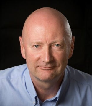This image shows a bald white man in a light blue shirt, smiling against a dark background. David is committed to accessible, inclusive banking.