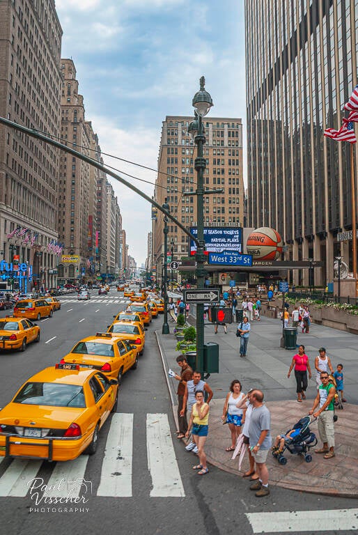 Yellow taxis line West 33rd Street near Madison Square Garden as pedestrians cross between tall buildings and street signs.