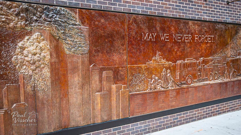 Bronze mural showing the Twin Towers, smoke, fire trucks, and firefighters, with the inscription “May We Never Forget” across the top.