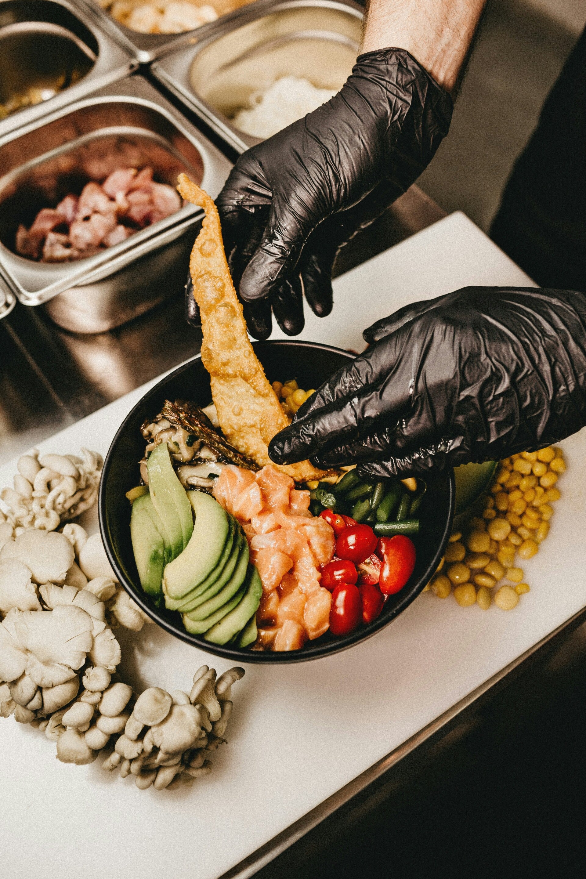 Food preparation of a salad bowl on a white counter top, by hands wearing black food safety gloves