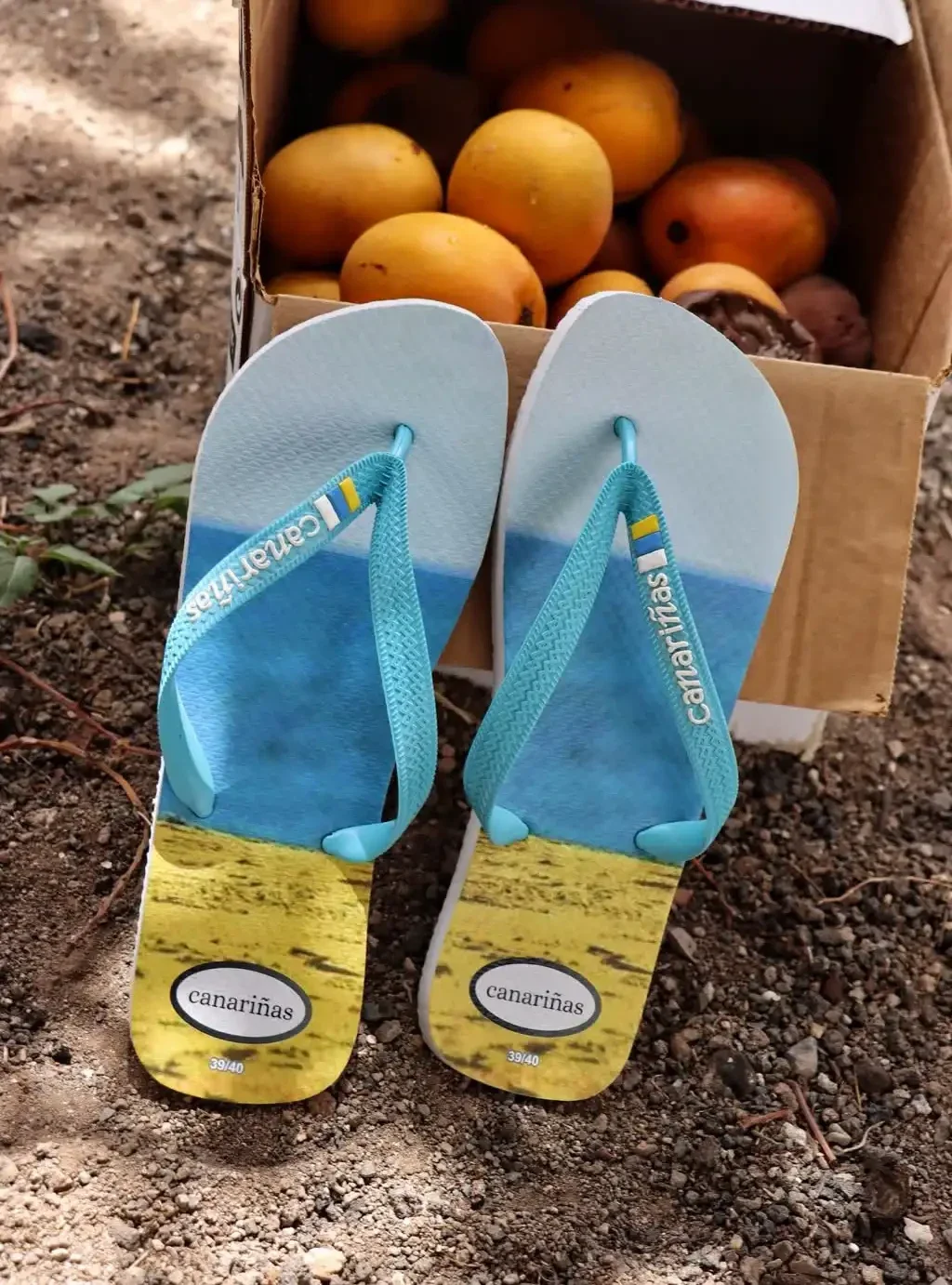 Chanclas de dedo Canariñas con diseño de bandera de Canarias junto a una caja de fruta tropical canaria.