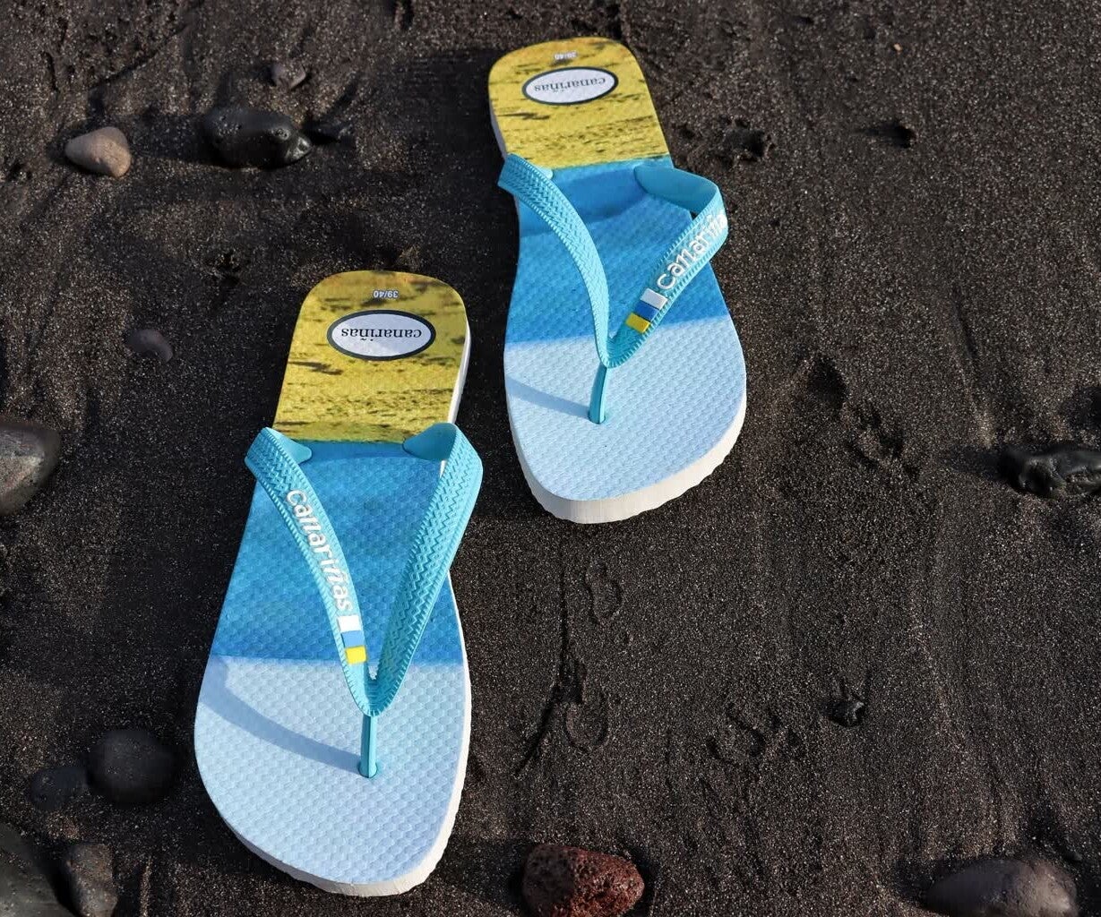 Chanclas de playa Canariñas sobre arena negra volcánica, calzado típico para vacaciones en las islas.