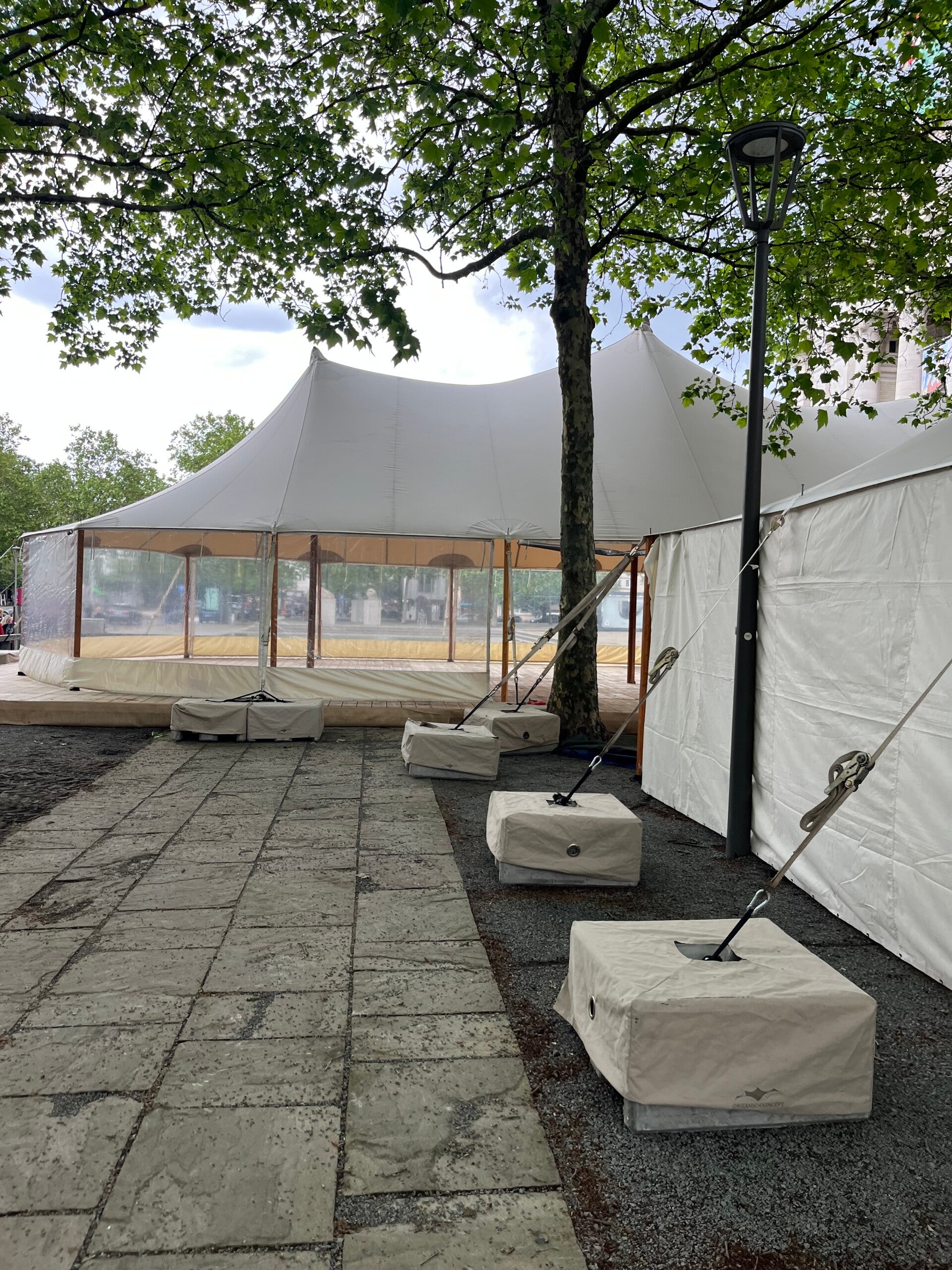 Silhouettetent in opbouw in Antwerpen met transparante zijwanden en ballastblokken voor verankering onder bomen.