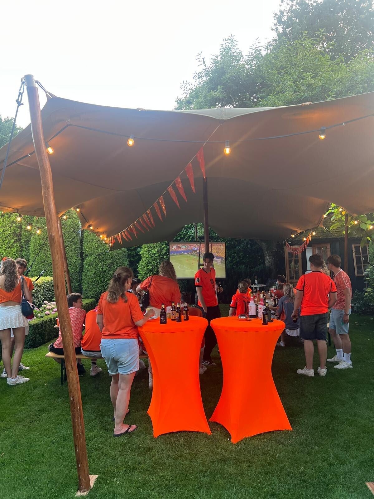 Tent versiert met oranje vlaggetjes en beeldscherm met oranje fans onder de tent