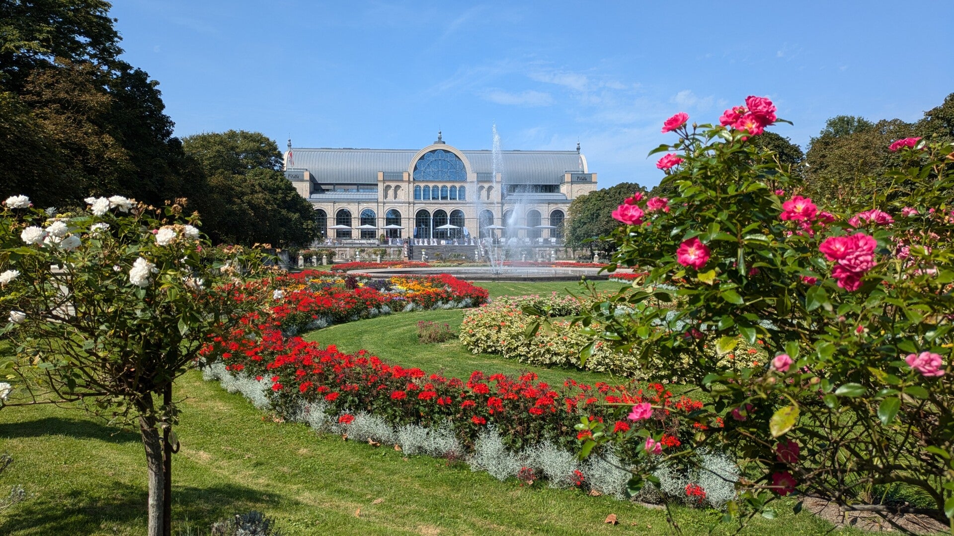 Blick auf das Restaurant Dank Augusta in der Kölner Flora, umgeben von bunten Blumenbeeten und üppigem Grün im botanischen Garten