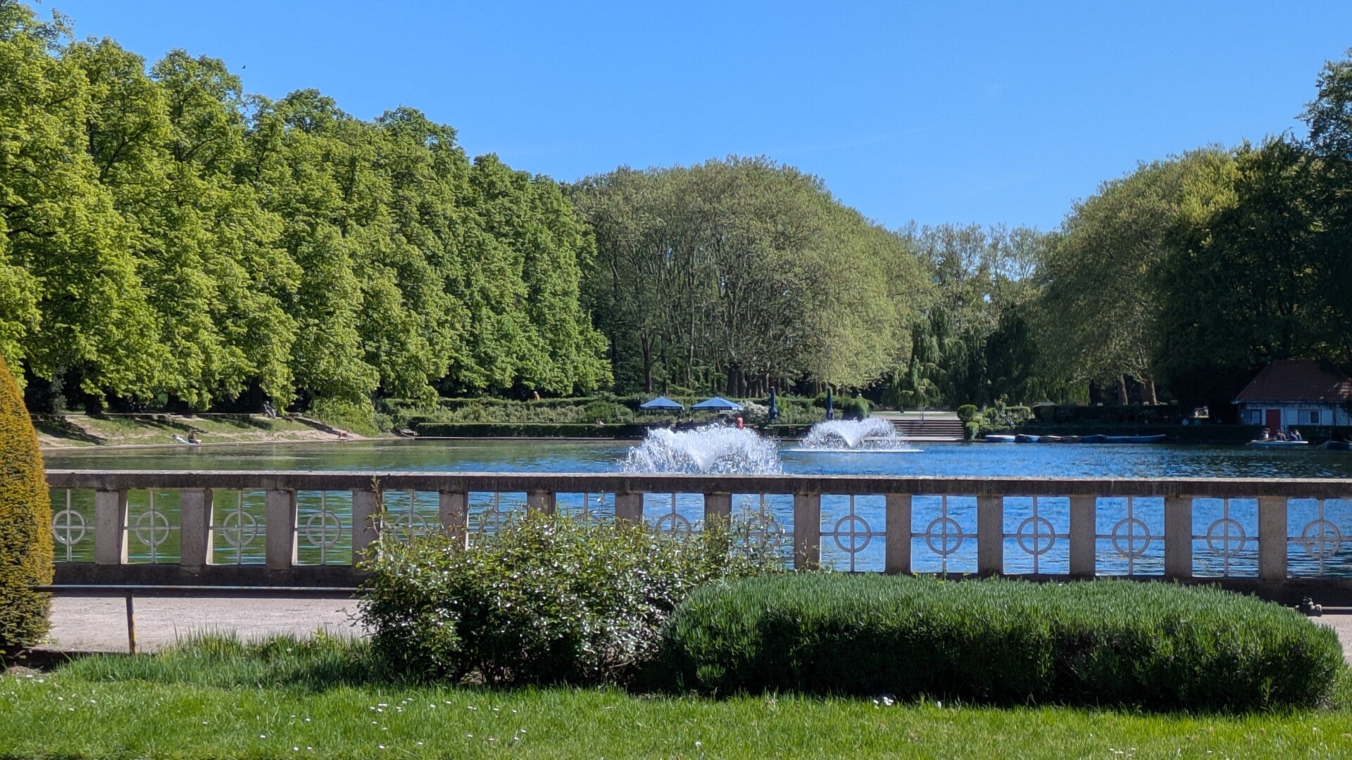Blick von einer grünen Wiese aus auf den Weiher mit zwei kleinen Wasserfontainen