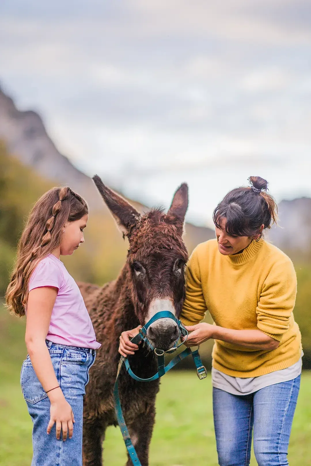 Séance de formation à la conduite et au travail en main d’un jeune âne, où une adulte encadre une enfant pour apprendre les gestes techniques de tenue du licol et de gestion de l’animal, dans un environnement naturel en montagne.