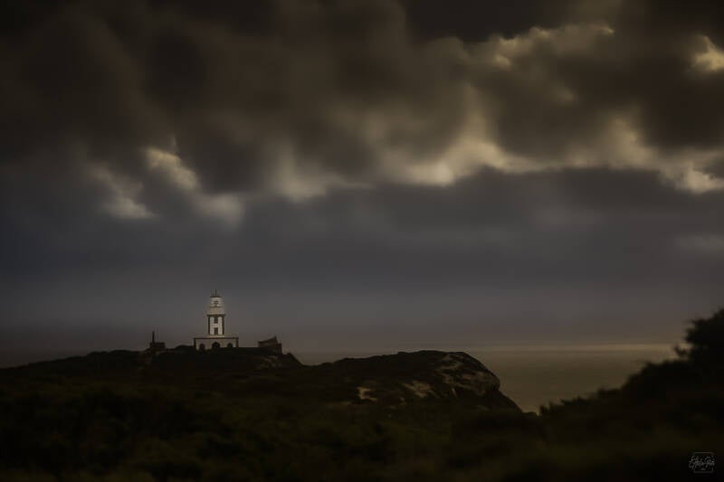 Phare solitaire immobile sur une falaise sombre, dominant la mer, sous un ciel dense et tourmenté où une lumière diffuse émerge des nuages.