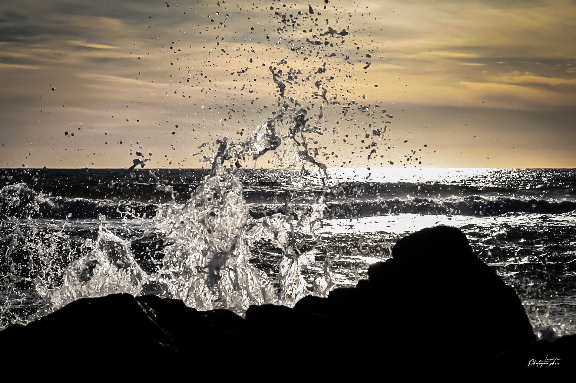 Éclaboussure de vague en contre-jour au coucher du soleil, eau figée en éclats lumineux devant les rochers