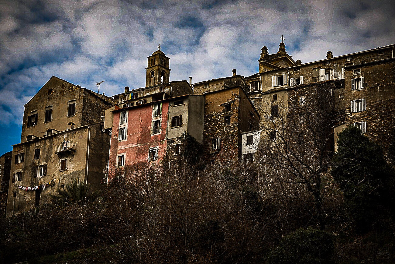 Village corse ancien sous un ciel lourd, façades de pierre chauffées par une lumière dramatique