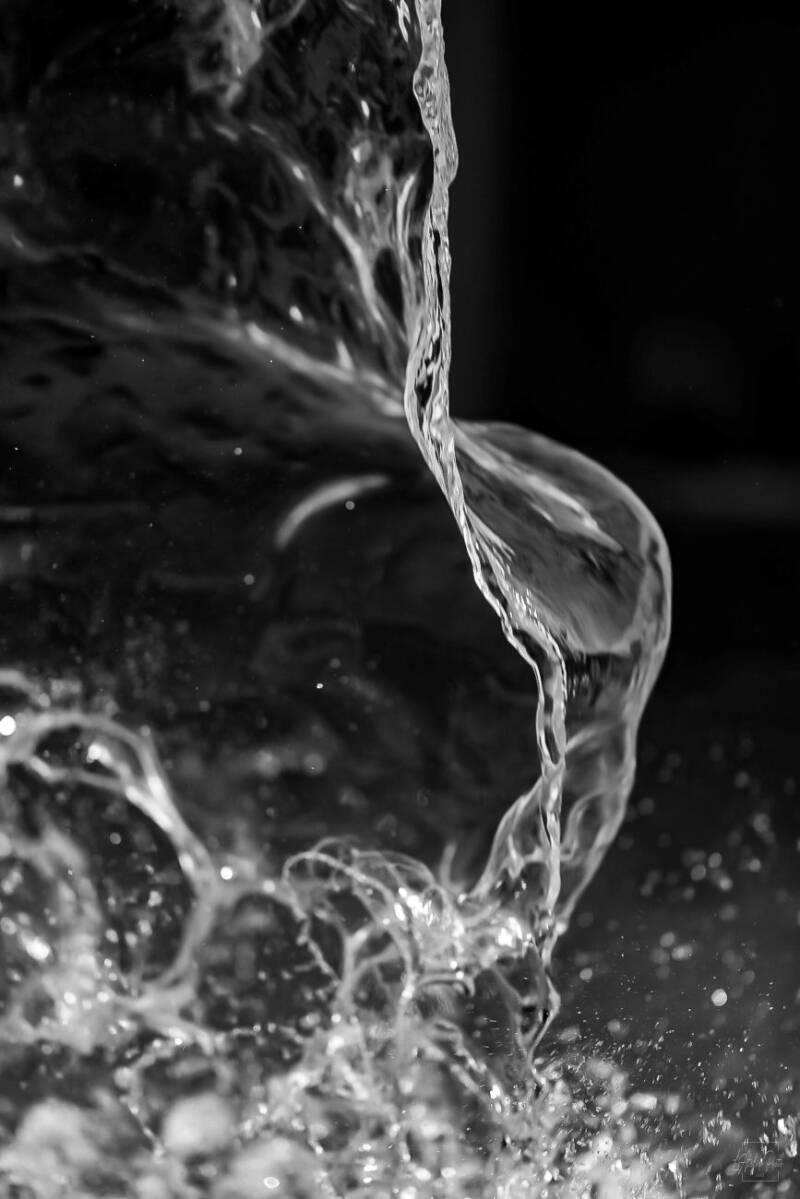 Macro en noir et blanc d’un jet d’eau courbe avec éclats lumineux.