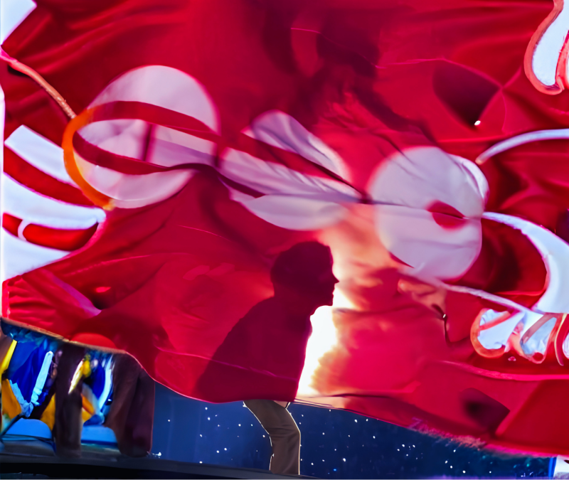 Imagen artística con una enorme tela roja translúcida que cubre el escenario. En el centro, se recorta la silueta oscura de un miembro de perfil, iluminado por un potente foco blanco posterior que crea un efecto místico. La tela tiene círculos claros proy