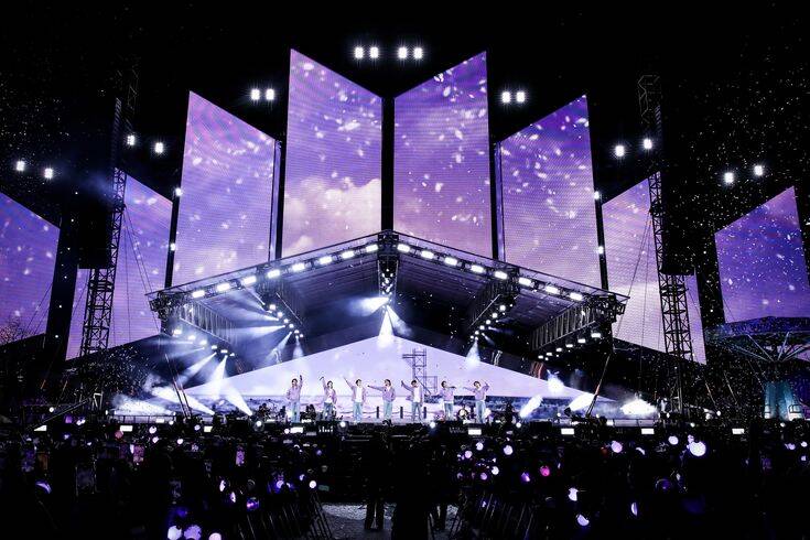 Concierto de BTS en un gran escenario iluminado con pantallas y luces moradas, frente a miles de fans ARMY con lightsticks encendidos, creando una atmósfera de unidad y emoción.