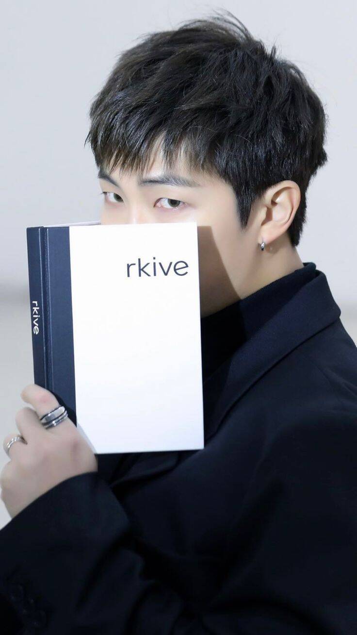 RM de BTS sosteniendo un libro que dice rkive, su cuenta personal en Instagram. Con el libro  oculta parte de su rostro, dejando solo al descubierto sus ojos.