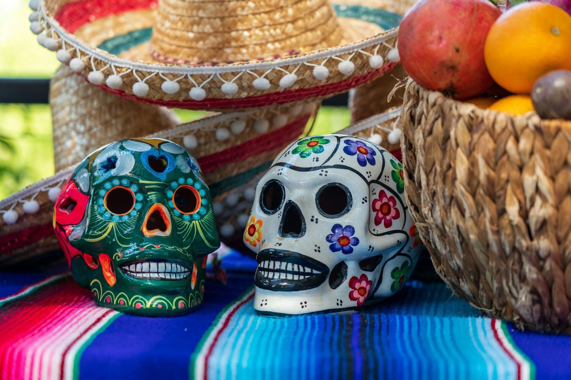Una mesa con dos calaveras de talavera, una canasta de fruta y sombreros regionales