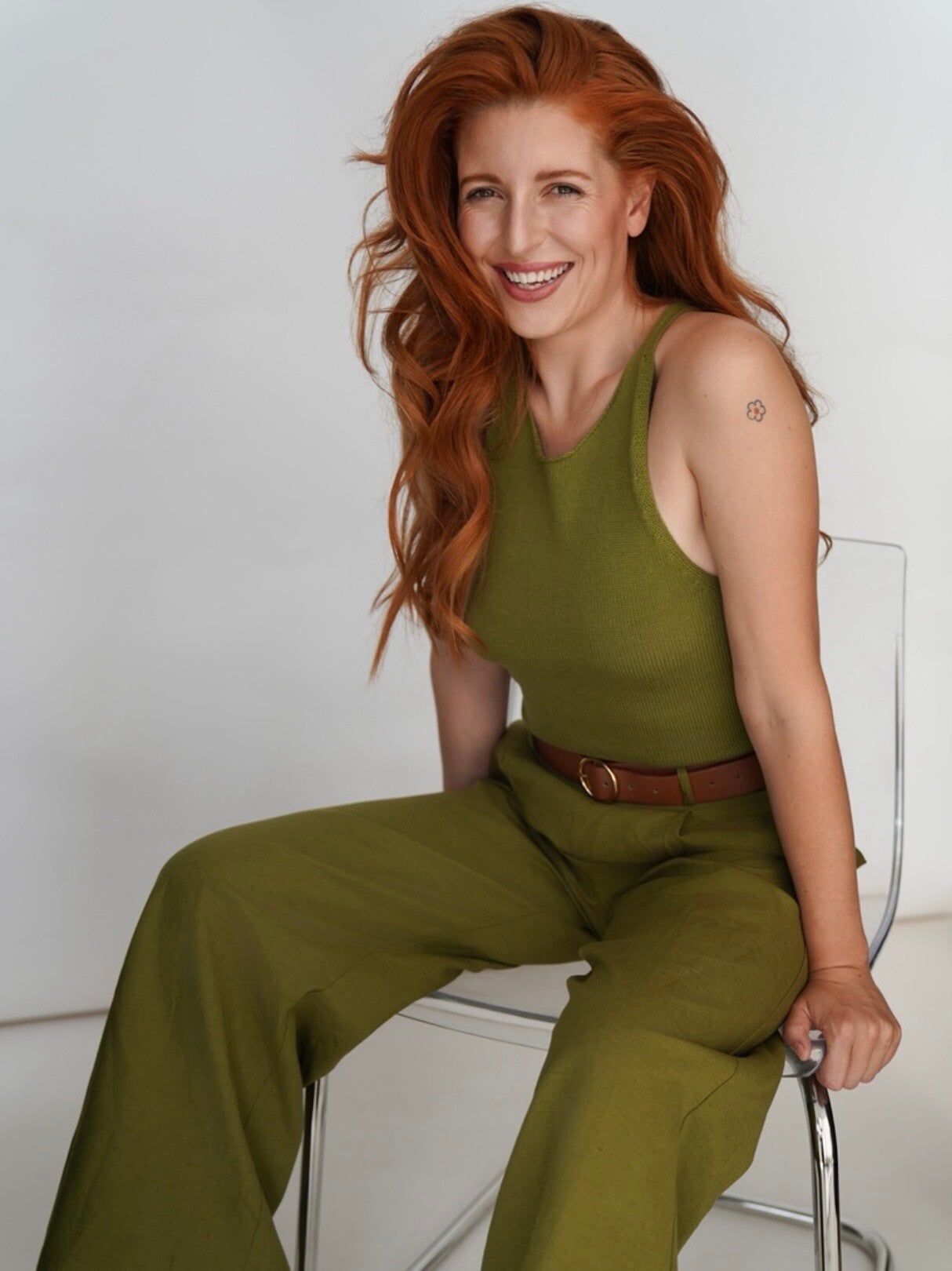 Bold red-haired woman gazing directly at the camera, dressed in olive green, featuring a subtle flower tattoo on her upper arm, showcasing a confident attitude.