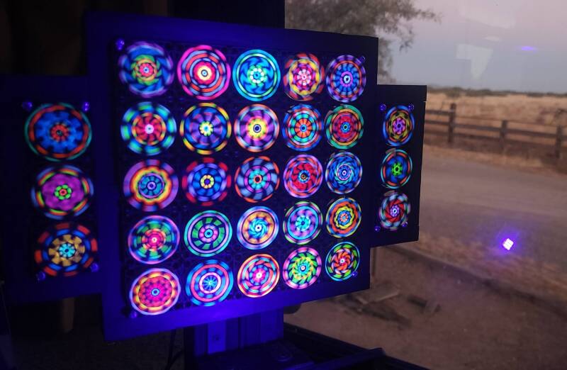 UV Blacklight Reactive Kinetic Micro Computer Fan Matrix with Colorful Hand-Painted Fluorescent Geometric Rotating Mandalas Shown Next to a Sunlit Daytime Window.