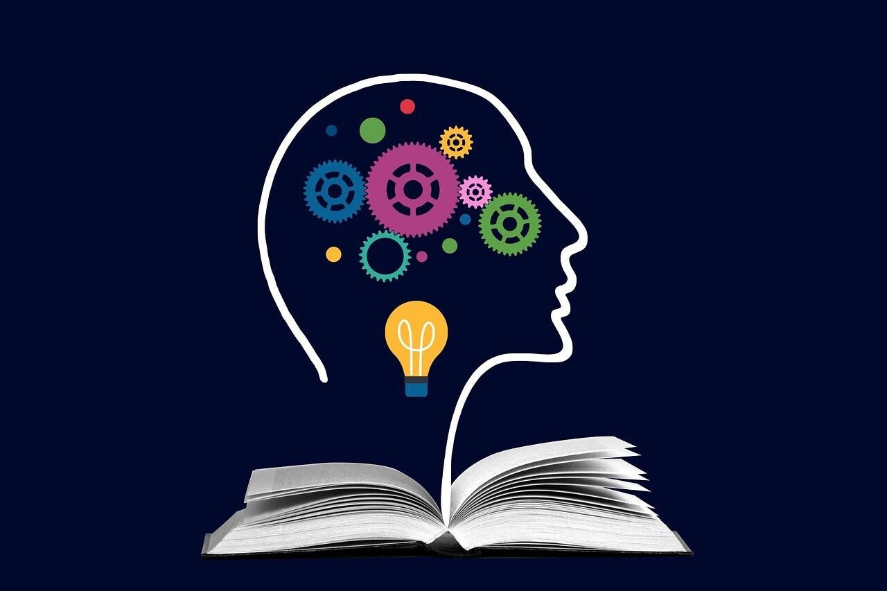 Offenes Buch mit einer stilisierten Kopf-Silhouette, aus der bunte Zahnräder und eine Glühbirne herausragen. Symbolisiert Lernen und Kreativität, Kontext Blog Jenseitskontakte Jana Lorenz.