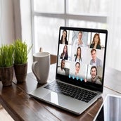 Laptop auf Holztisch zeigt neun Personen in einem Videoanruf, daneben zwei Pflanzen in Töpfen und eine weiße Kaffeetasse, im Kontext der Großen Gruppenlesung mit Medium Jana Lorenz.