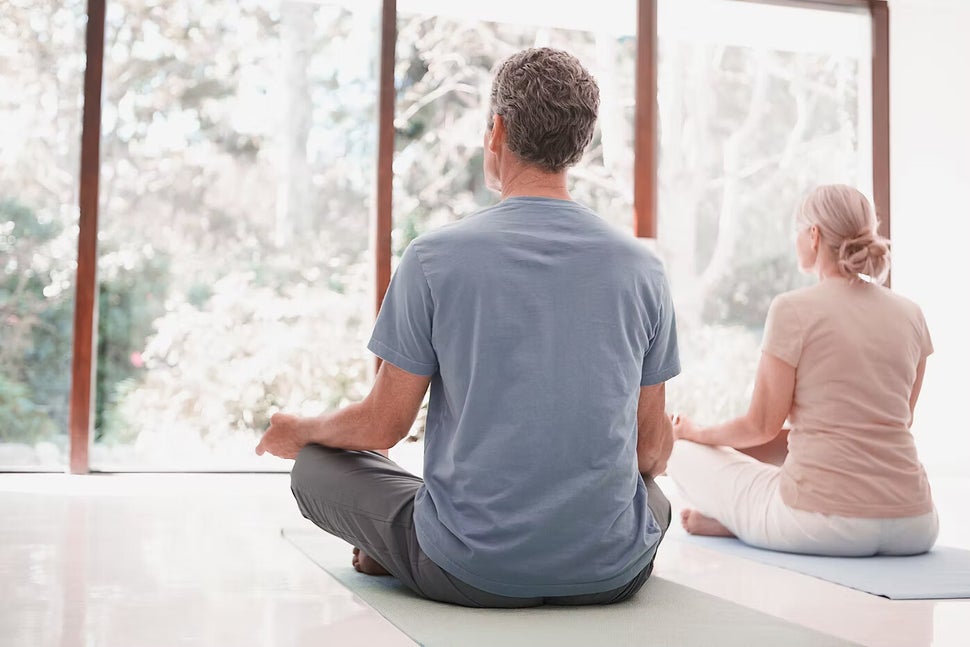 Zwei Personen sitzen im Schneidersitz auf Yogamatten. Ein Mann mit grauem T-Shirt und eine Frau mit beigem Oberteil meditieren in einem hellen Raum mit großen Fenstern, Kontext Blog Jenseitskontakte Jana Lorenz.