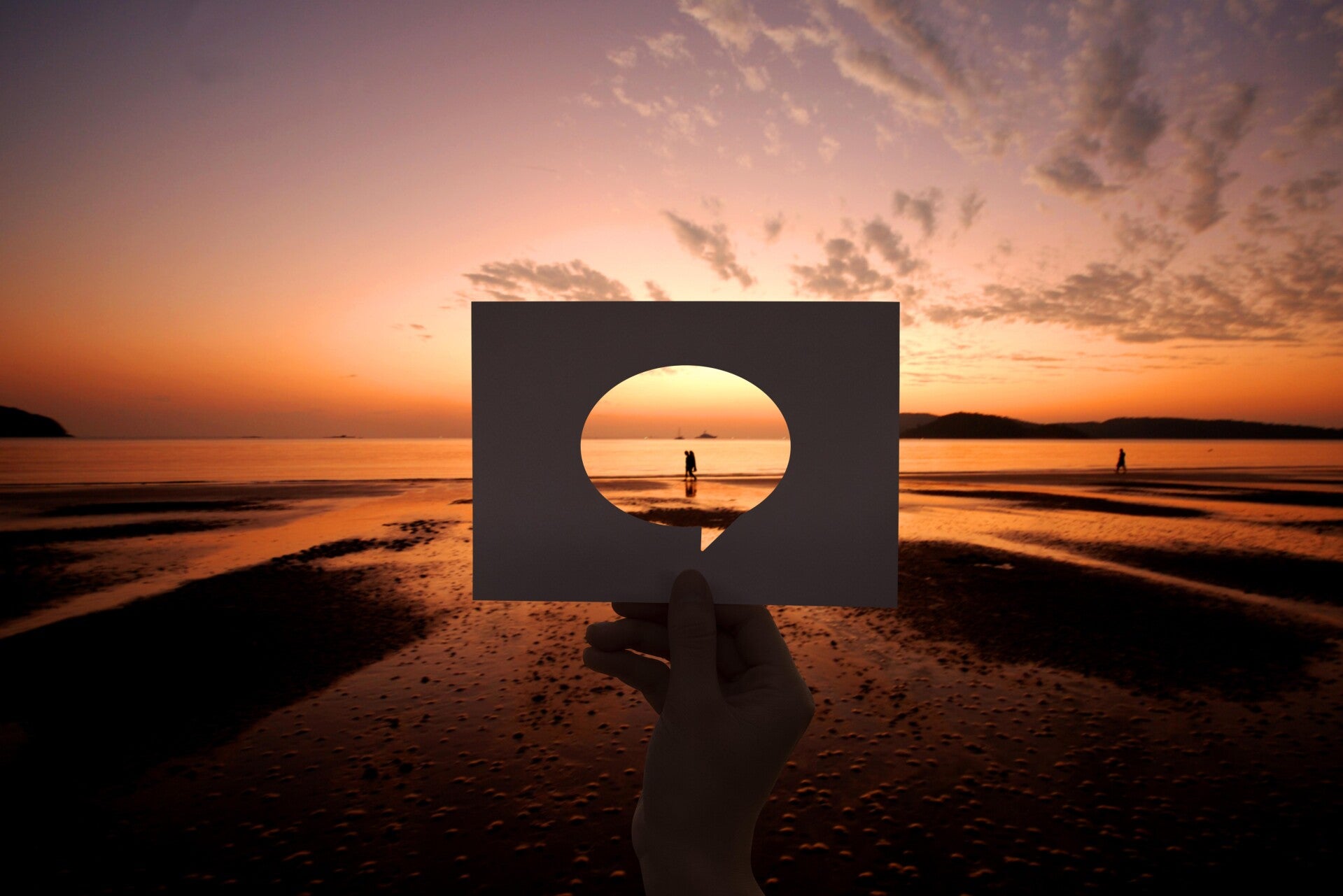 Hand hält rechteckiges Papier mit ovalem Loch vor Sonnenuntergang am Strand, Person im Hintergrund sichtbar, Kontext Blog Jenseitskontakte Jana Lorenz.