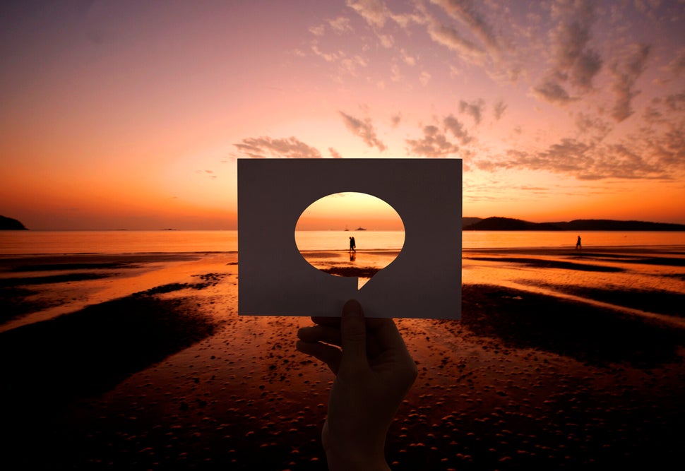 Hand hält rechteckiges Papier mit ovalem Loch vor Sonnenuntergang am Strand, Person im Hintergrund sichtbar, Kontext Blog Jenseitskontakte Jana Lorenz.