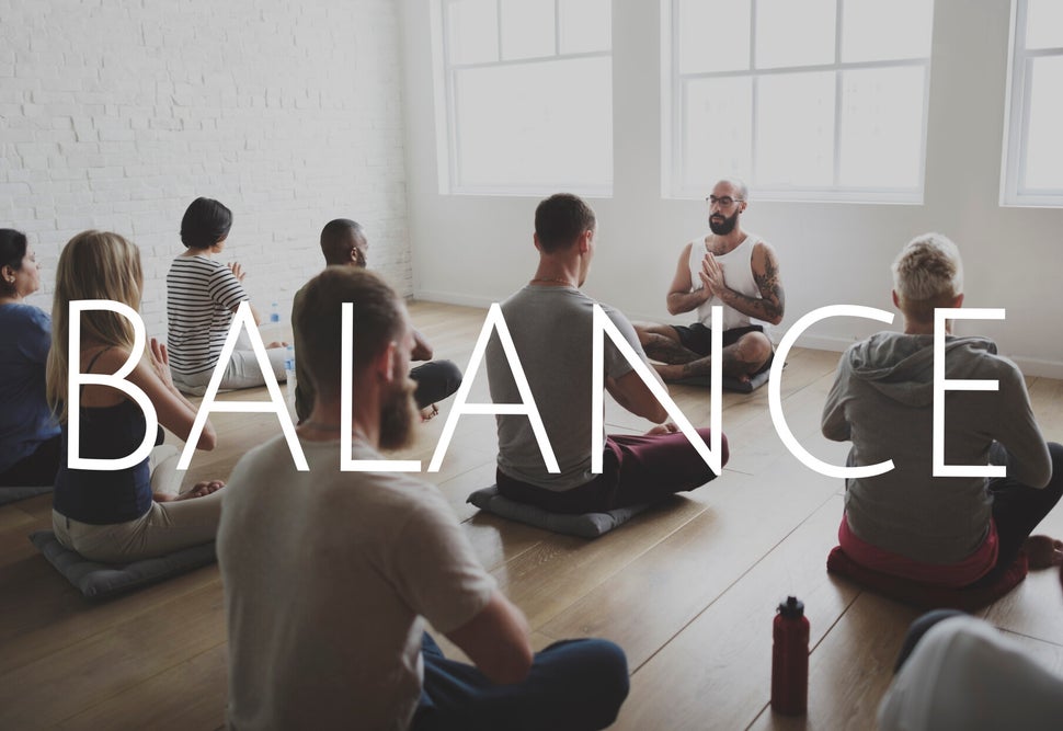 Gruppe von acht Menschen sitzt im Schneidersitz auf Yogamatten in einem hellen Raum, vorne sitzt ein Mann mit tätowierten Armen und verschränkten Händen, darüber steht das Wort 'BALANCE'