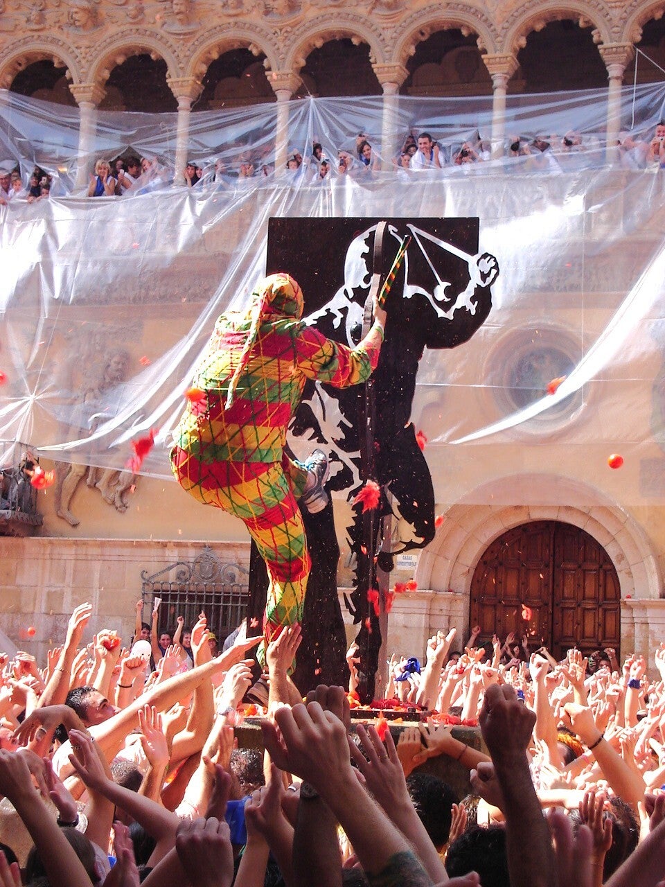 El Cipotegato, vestido de arlequín, avanza entre la multitud bajo una lluvia de tomates en Tarazona  (97 car.)  Encaja con la fiesta: el arlequín a tomatazos es justo la estampa. Es BY-SA 3.0, no 4.0. Foto de 2007, pero el rito es idéntico; vale.  Recorda