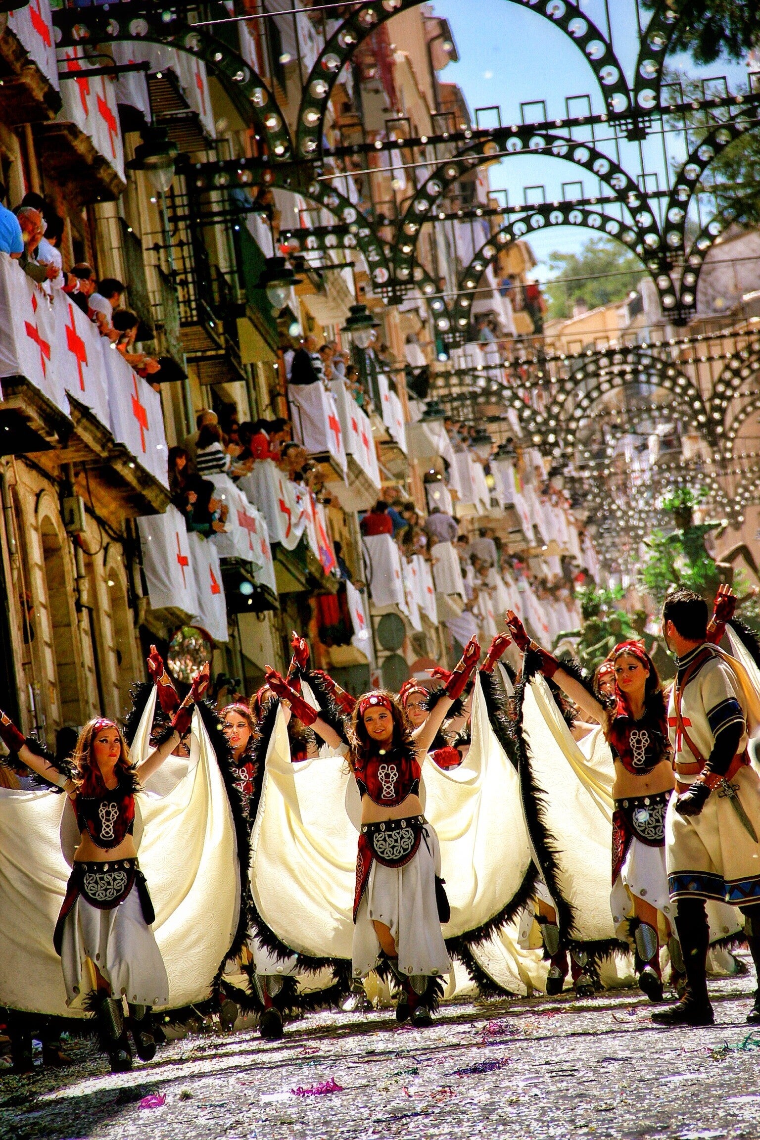 Bailarinas con grandes alas blancas desfilando por una calle de Alcoy engalanada con banderas de la cruz y la enramada, durante los Moros y Cristianos.