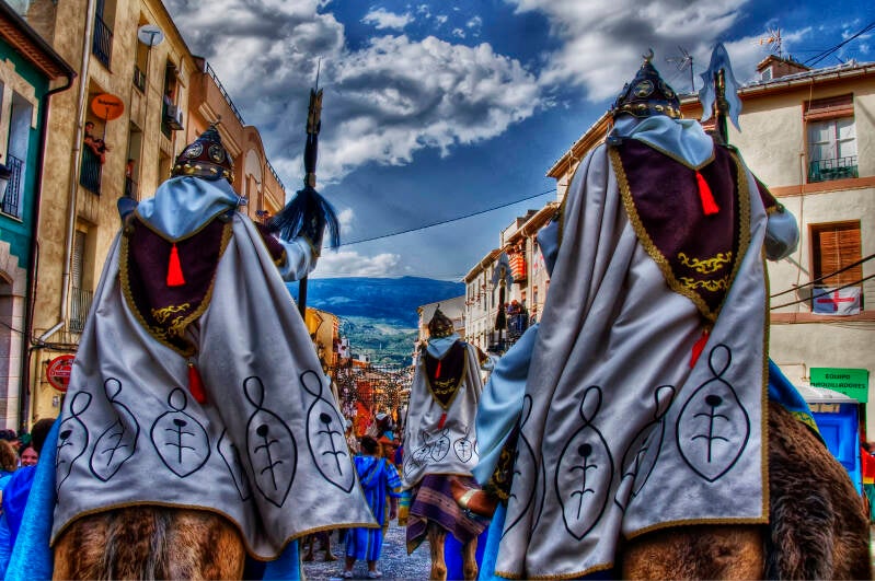 Jinetes de espaldas con capas bordadas desfilando por una calle de Alcoy, con las montañas al fondo, durante los Moros y Cristianos.