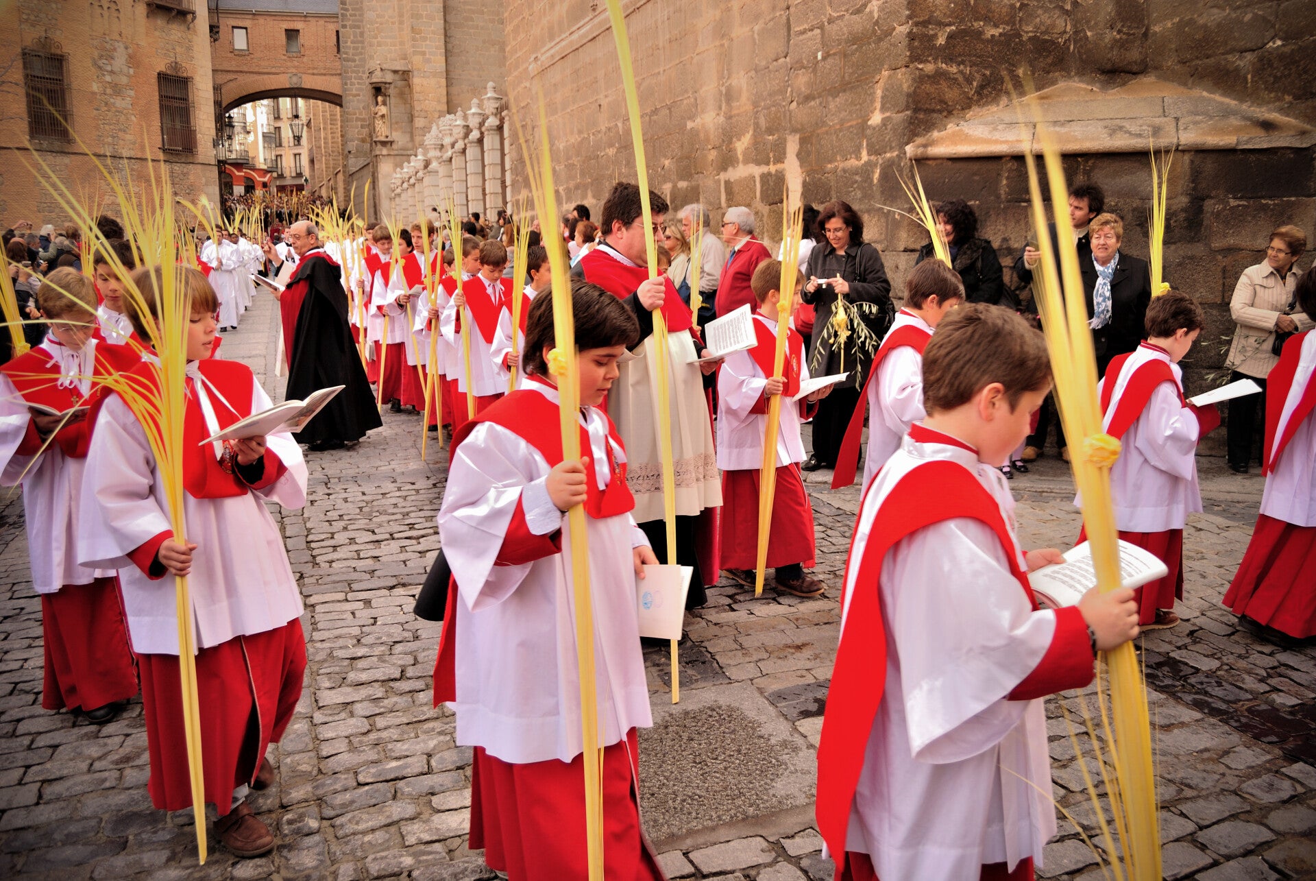 Monaguillos con palmas y cofrades recorren una calle empedrada de Toledo en la procesión del Domingo de Ramos