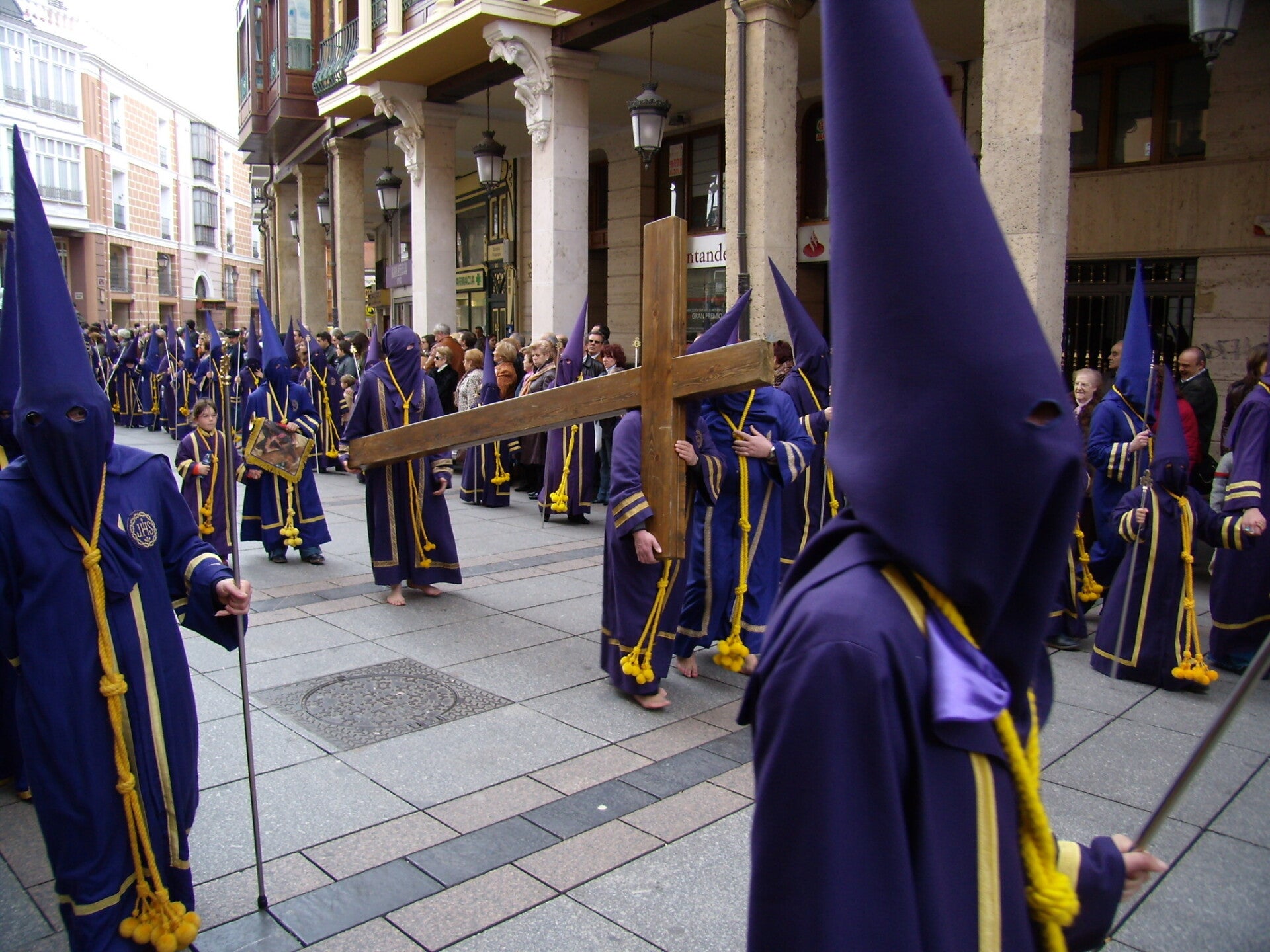Nazarenos con túnica y capirote morados y cordón amarillo, uno de ellos con una cruz de madera, en la procesión de los Pasos del Viernes Santo en Palencia.