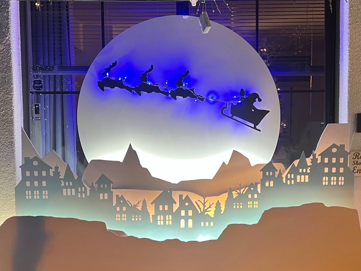 A magical winter scene features Santa Claus and his reindeer silhouette flying across a full moon, hovering over a snowy city with rooftops, chimneys, and glowing lights