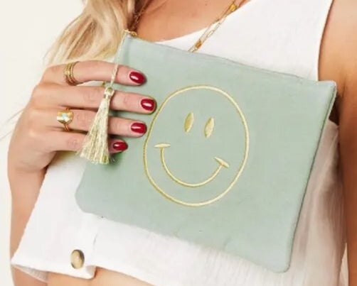 Eine mintfarbende Kosmetiktasche mit einem goldenen Smiley wird von einer Frauenhand gehalten.