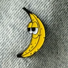 Ein Ansteckpin in Form einer Banane mit einem Gesicht steckt an einem jeansartigen Stoff. Die banane hat großen Augen und lächelt.
