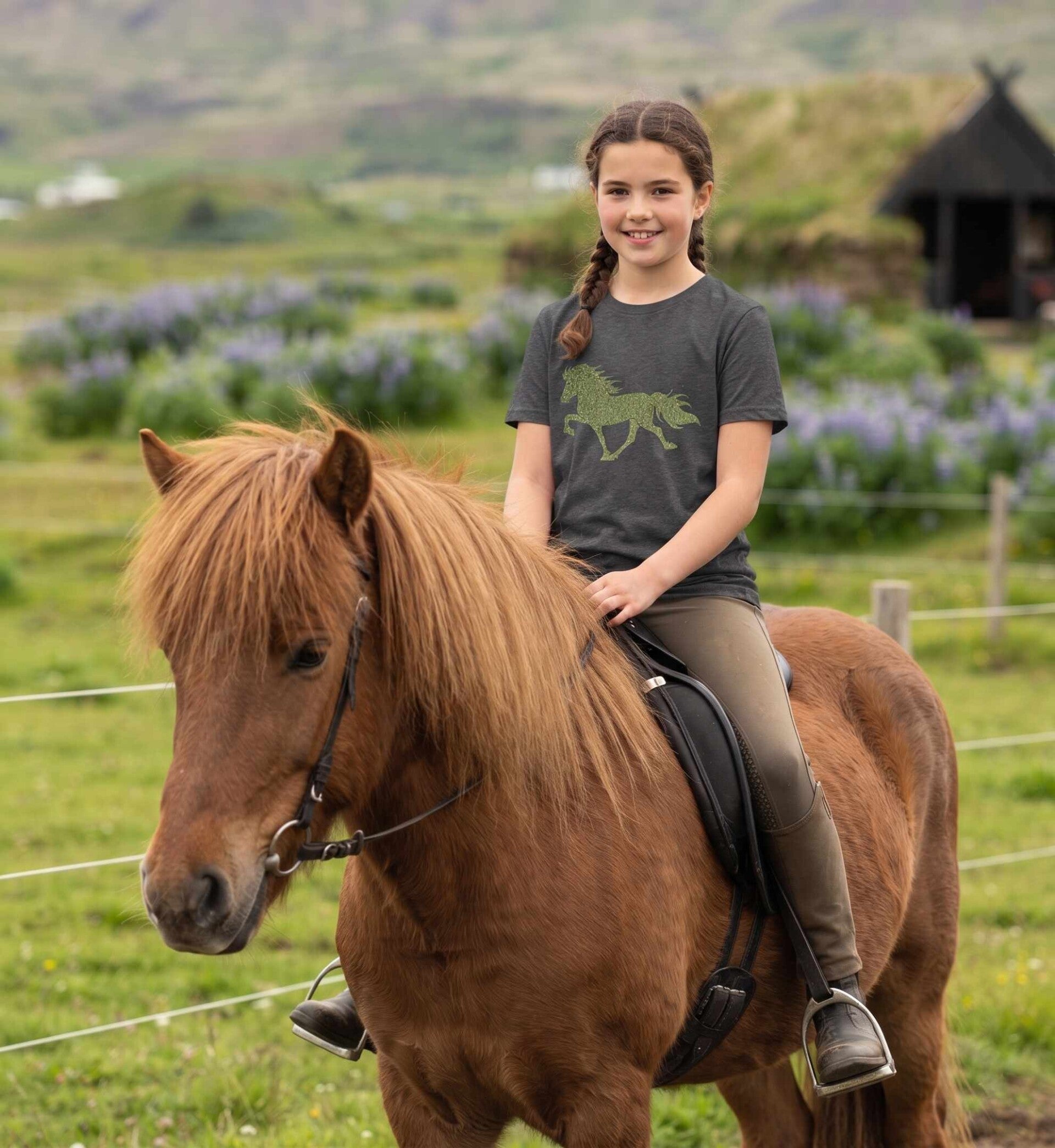 Ein ca. 12-jähriges dunkelhaariges Mädchen mit Zopffrisur sitzt auf einem fuchsfarbenen Islandpferd . Sie trägt ein anthrazitfarbenes Shirt mit einem  Tölter-Bügelmotiv in grünem Glitzer. Standort: Island